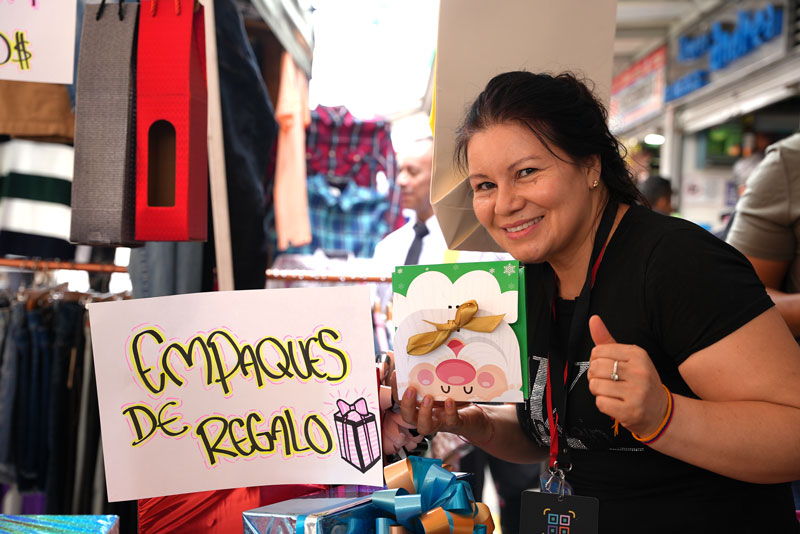 Mujer sonriente mostrando un empaque de regalo temático, junto a un letrero que dice -Empaques de Regalo- en un puesto comercial